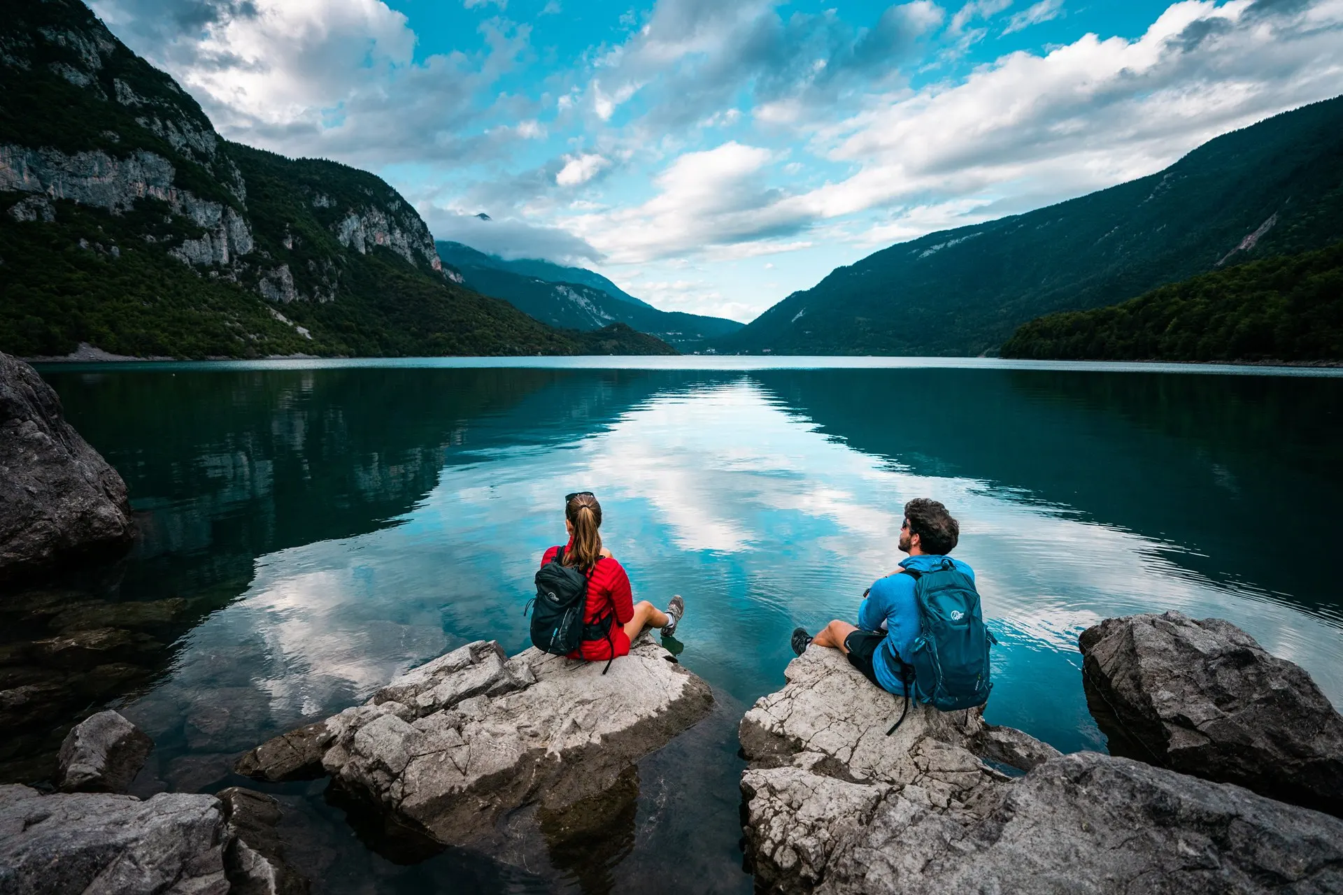 Two hikers sitting on the rocks by Lake Molveno, with the mountains reflected in the water
